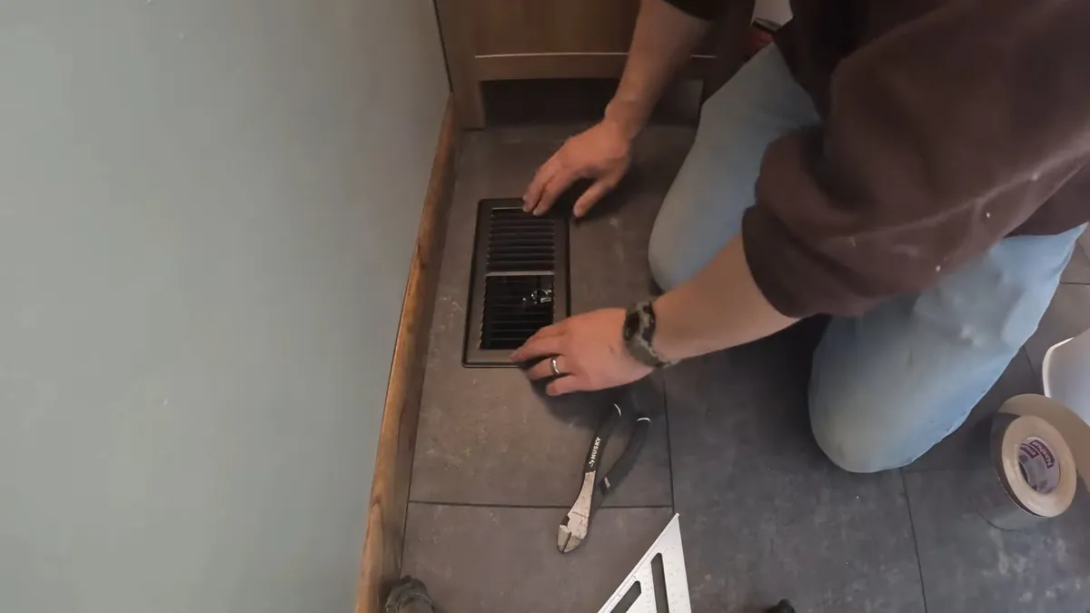 Person kneeling beside an open floor drain opening in a tiled floor with pliers and tape nearby