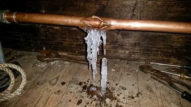 Copper pipe split along its length with long icicles hanging from the break above a wooden floor