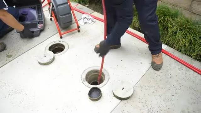 Technician feeding a red drain cable into an open cleanout on a concrete slab, with a camera cart beside them