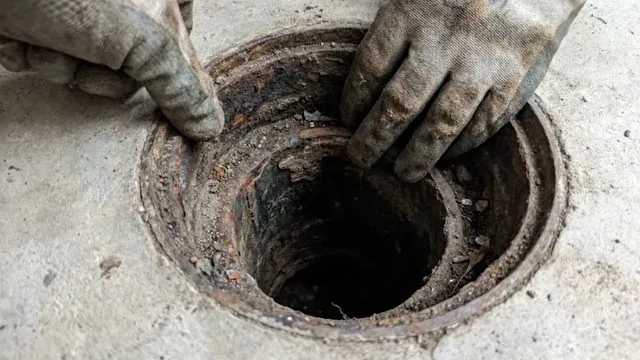 Gloved hands at the rim of a rusted round outdoor drain set in a concrete slab