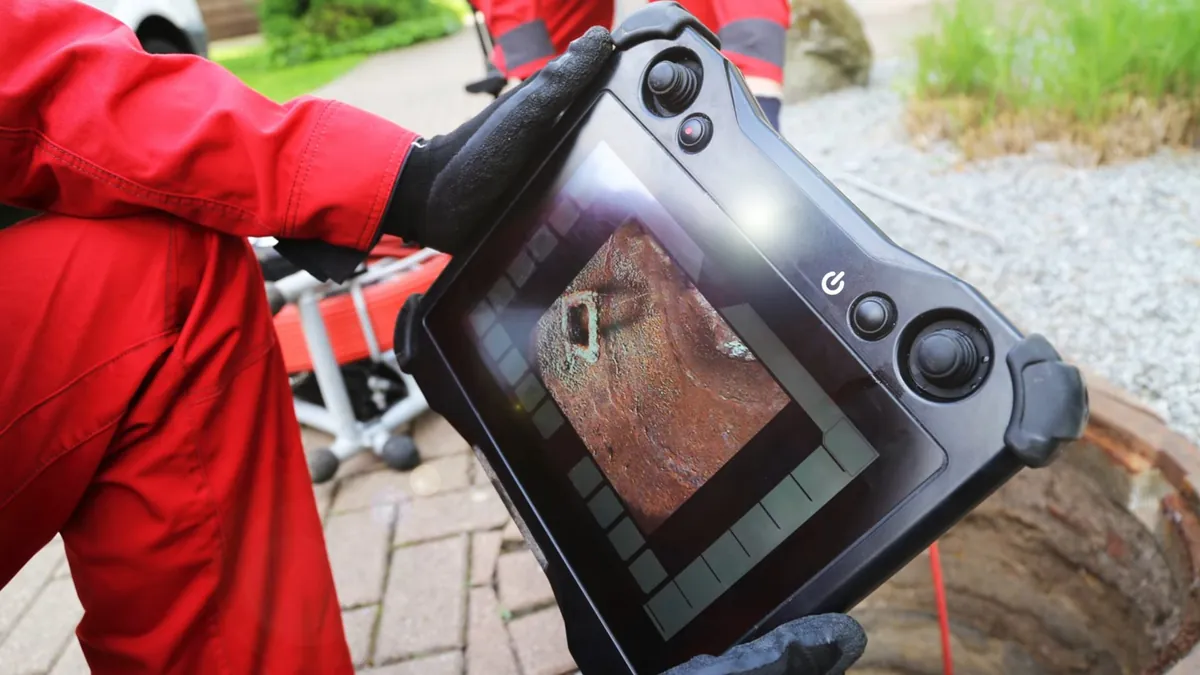 Technician in a red jacket holding a handheld pipe camera monitor that shows the inside of a line