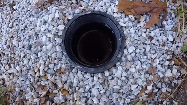 Open black cleanout pipe set in white gravel with fallen leaves around it