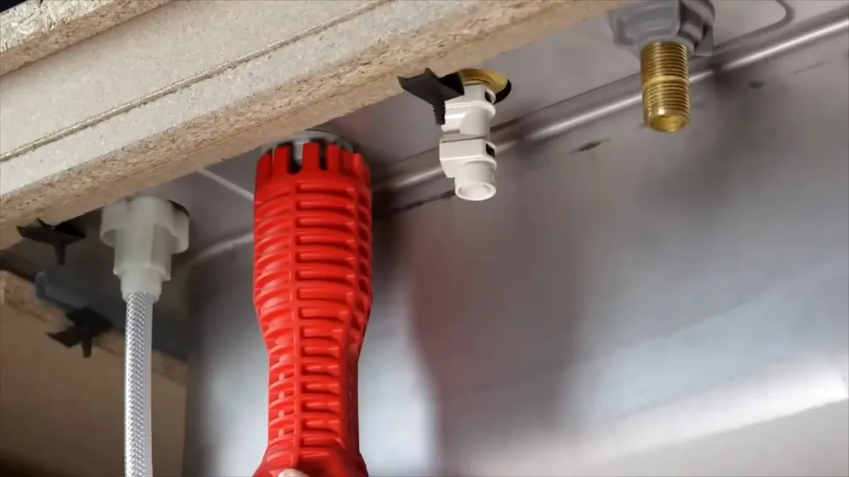 Red basin wrench held under a sink beside a white supply stop and a brass fitting