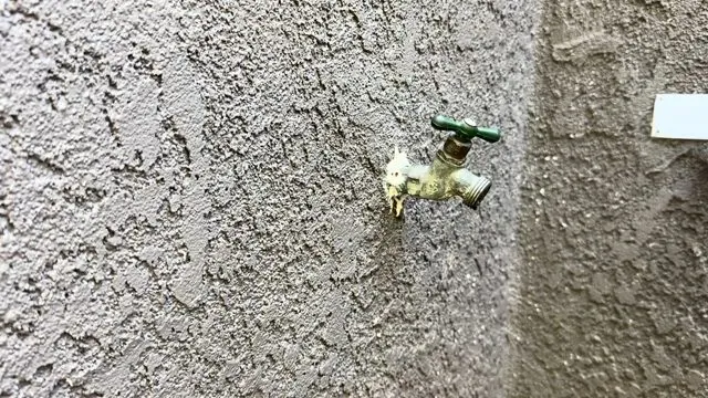 Brass hose bib with a green handle on a grey stucco exterior wall