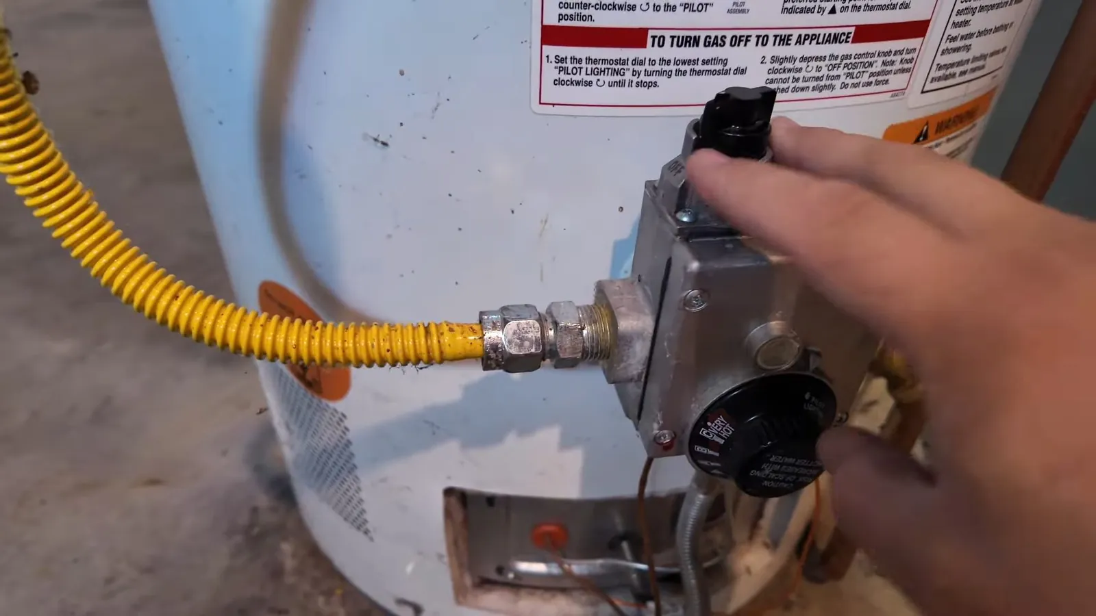 Hand turning the gas control on a water heater beside a yellow flexible gas connector