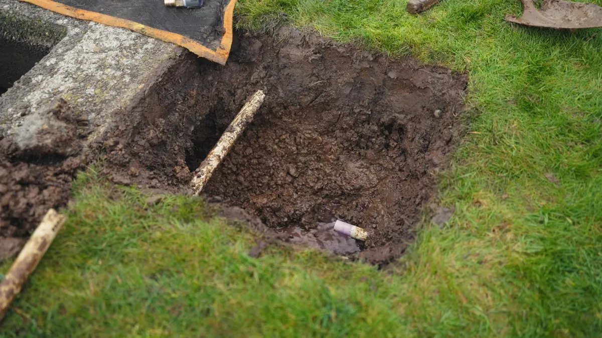Rectangular hole dug in a lawn beside a concrete edge with a wooden stake in it