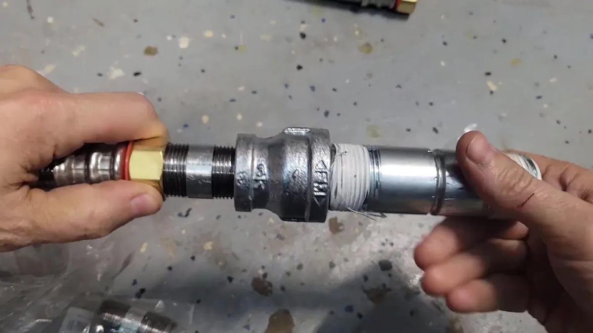 Hands joining a galvanised steel pipe coupling to a threaded brass fitting, with white thread tape on the pipe end