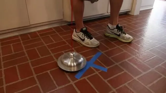 Metal listening disc on a brick-pattern tile floor with a blue tape cross beside it and a person standing behind