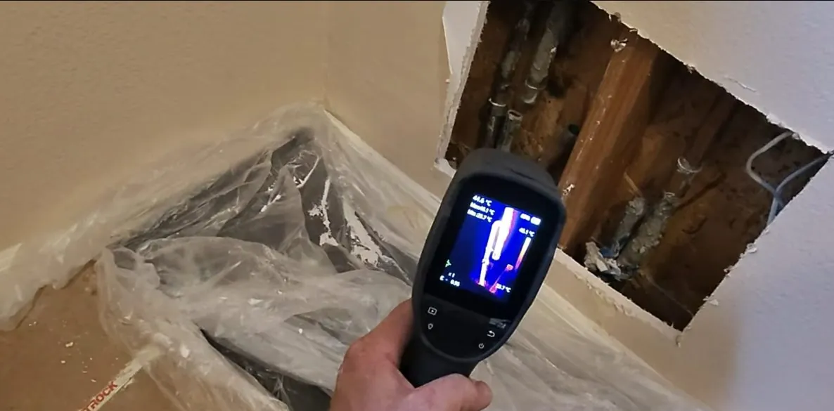 Handheld thermal camera showing a heat image beside an opened wall cavity