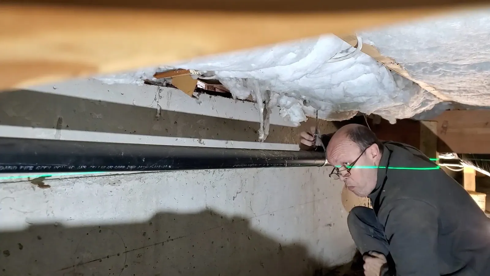 Man crouching in a crawlspace and looking along a black pipe under insulated floor joists