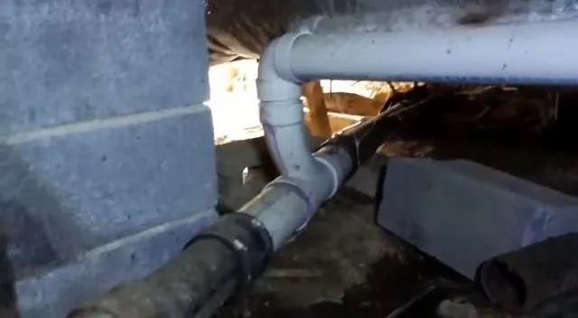 White PVC drain elbow and black waste pipe running under a house beside a block wall
