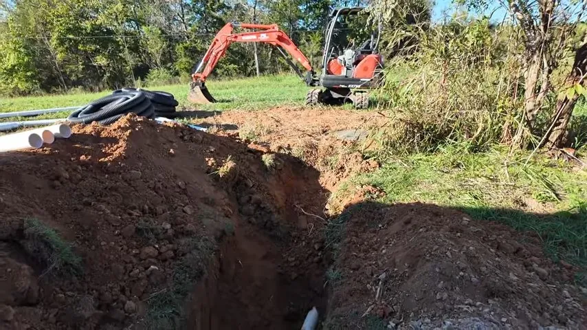 Small orange excavator beside a long open trench in a grassy field