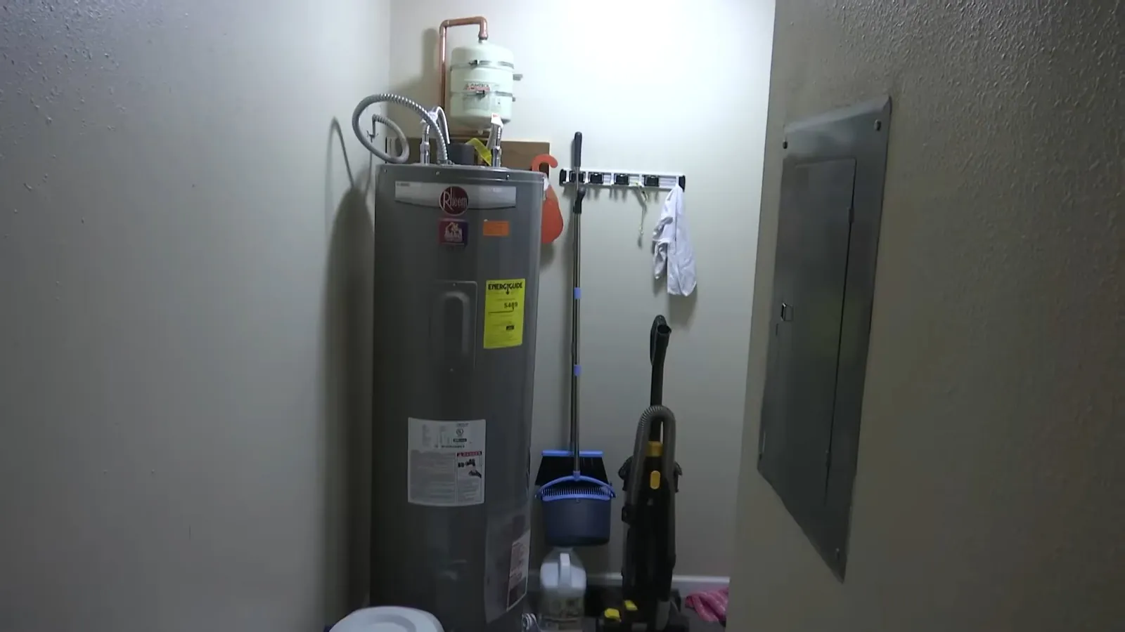 Electric water heater stood beside an electrical panel with brooms and a vacuum next to it