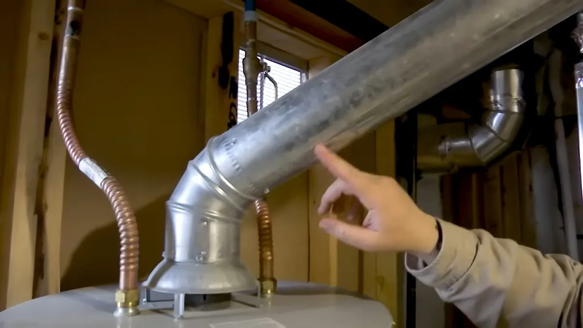 Hand pointing at the galvanised vent pipe on top of a water heater, with two flexible supply lines either side