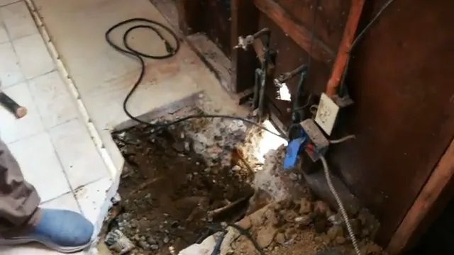 Hole broken through a tiled floor beside a wall with exposed pipes and a cable
