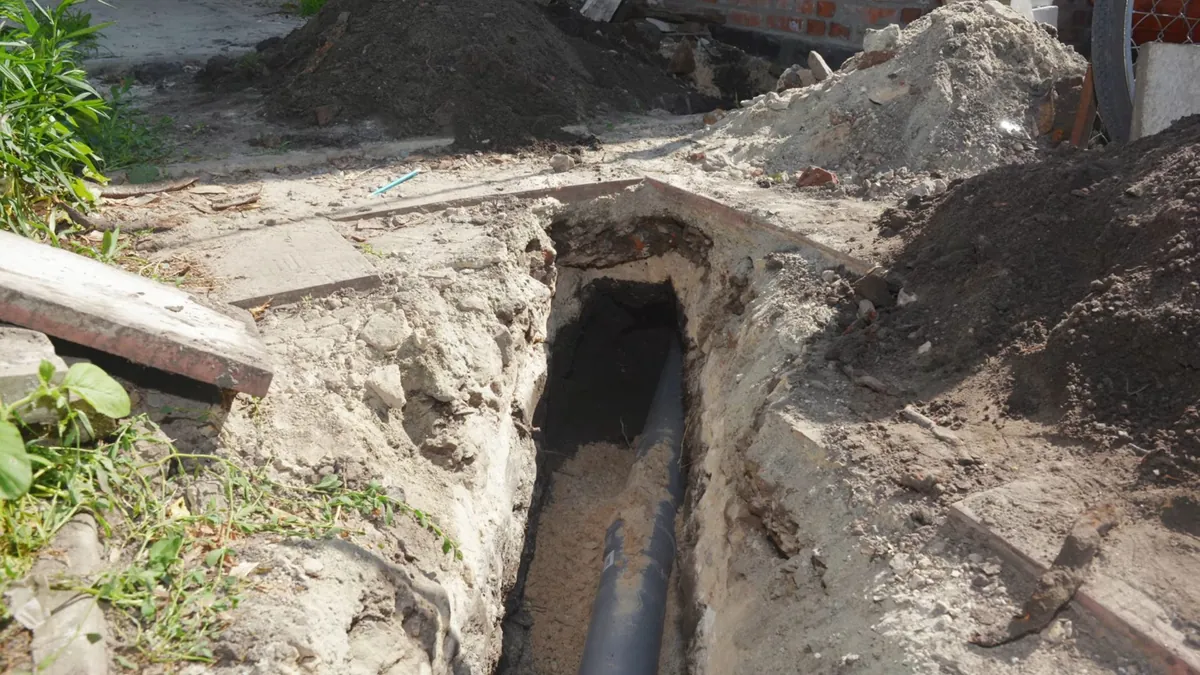 Narrow excavation beside a pavement slab with a dark pipe visible at the bottom