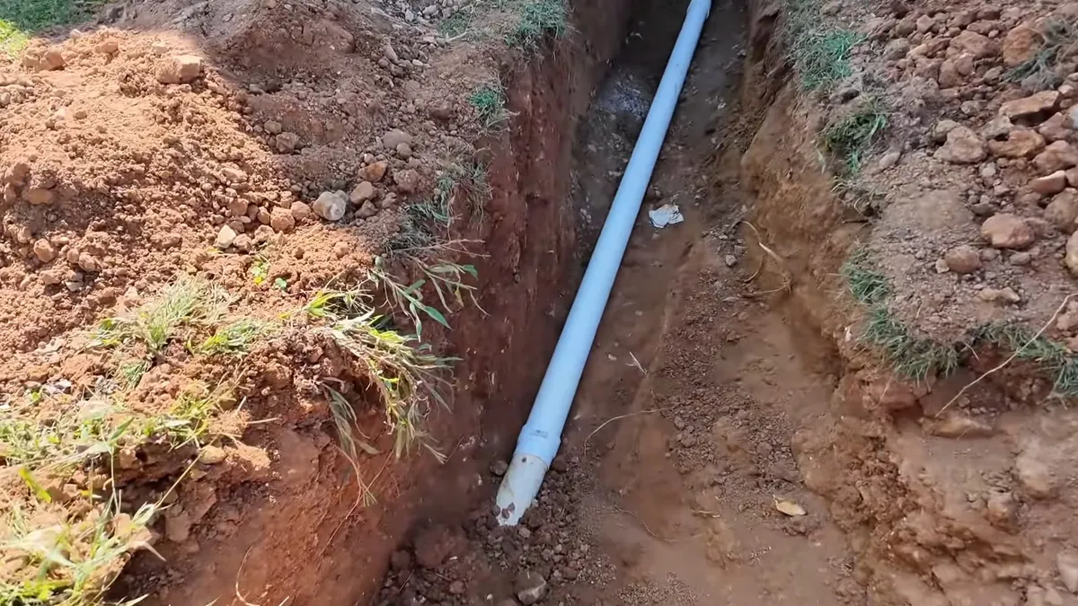 Narrow trench in red clay soil with a pale blue pipe laid along the bottom