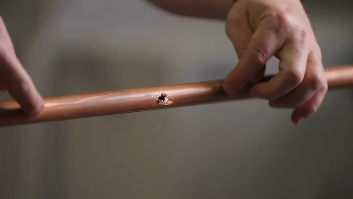 Two hands holding a length of copper pipe with a small pinhole in its wall
