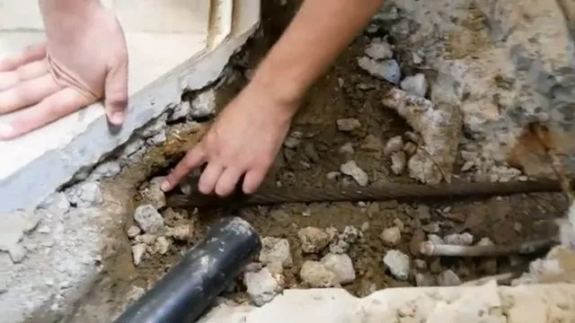 Hand resting on broken concrete beside a black drain pipe laid in soil beneath a slab edge