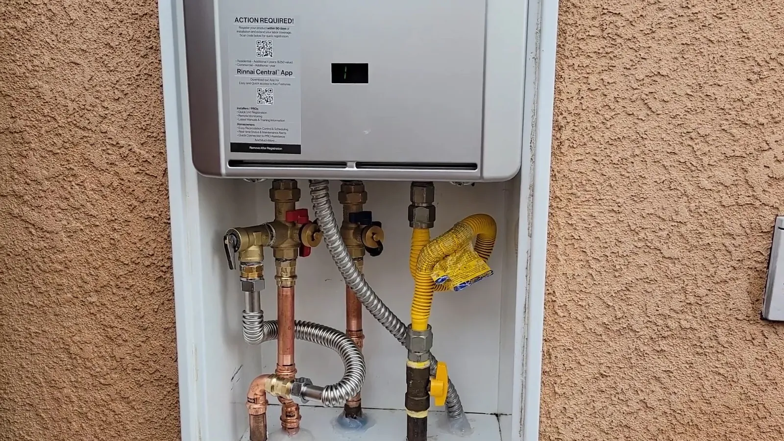 Tankless water heater in a recessed stucco wall box with copper pipes, braided hose and a yellow gas connector below