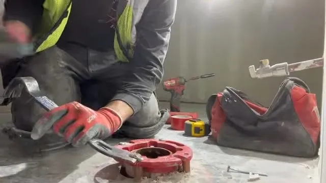 Plumber in a high-visibility vest kneeling at a red closet flange with a tool bag and tape measure nearby