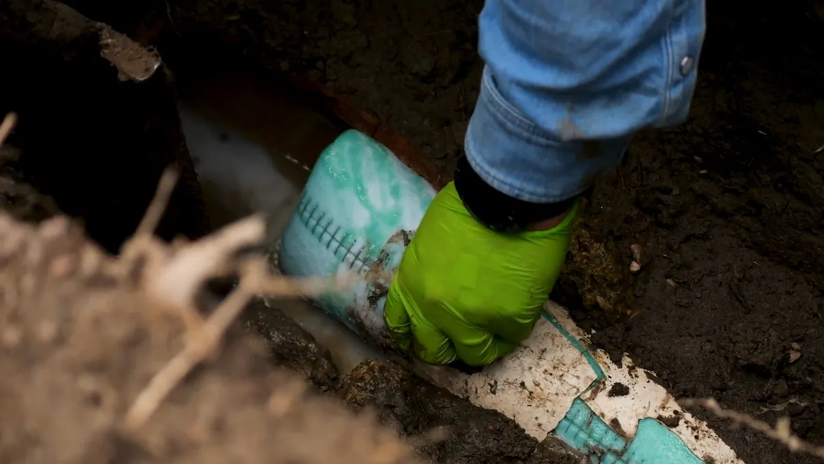 Hand in a green glove guiding a teal printed liner tube in a dug trench