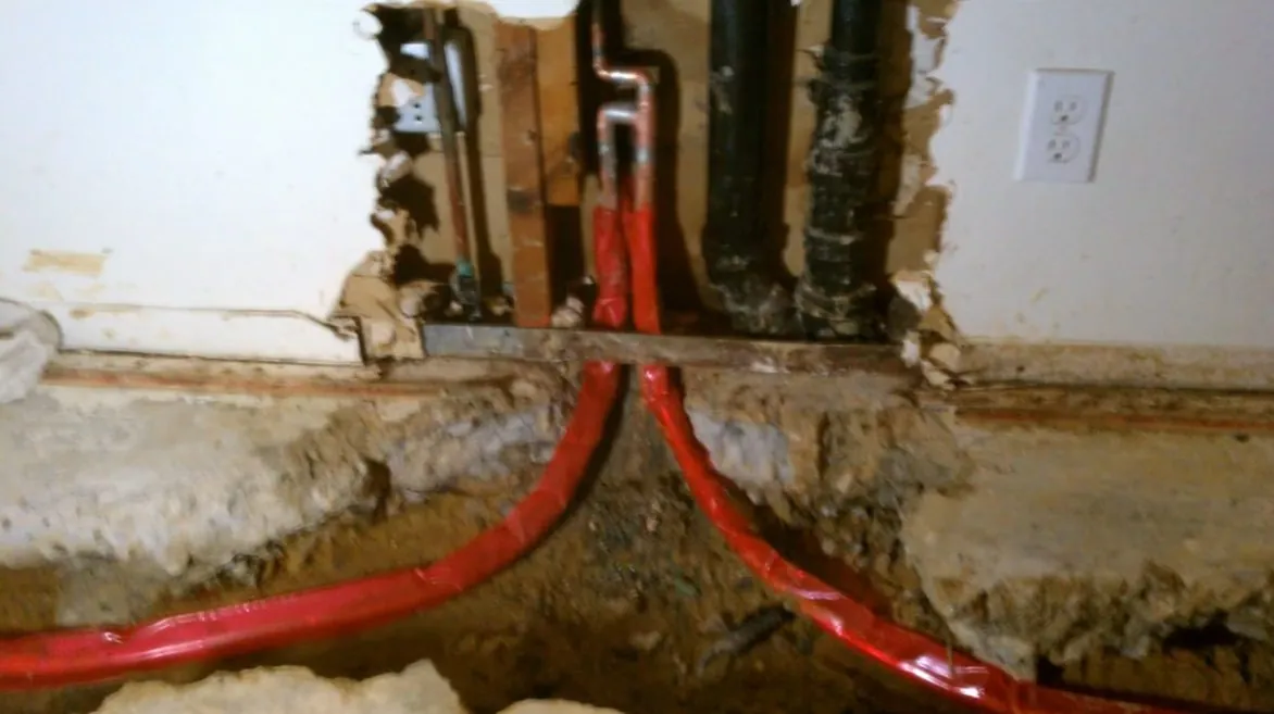 Base of a wall cut open to the slab showing red PEX lines and black pipe in the gap