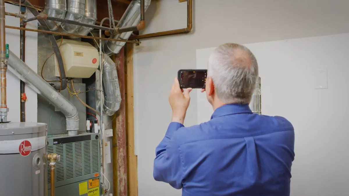 Man in a blue shirt photographing a water heater and its flue with a phone