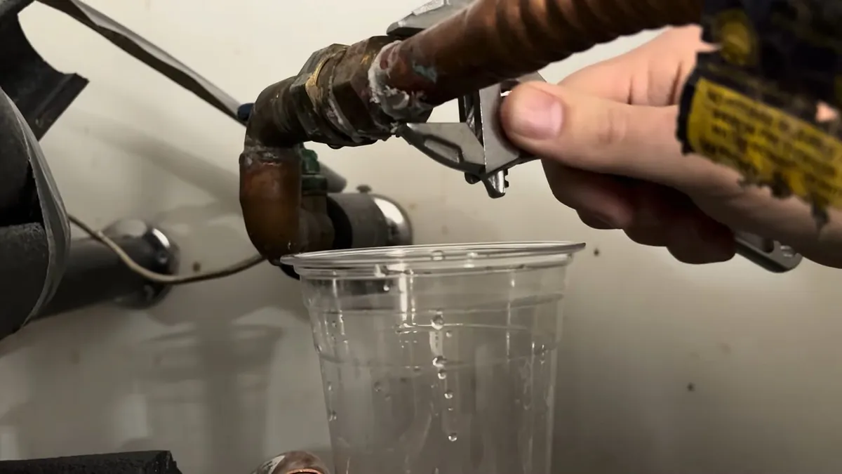 Hand with pliers at a heater drain valve running water into a clear plastic cup